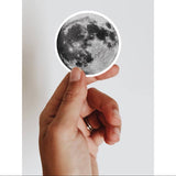 Hand holding a vinyl round full moon sticker, perfect for any lover of the moon or space!